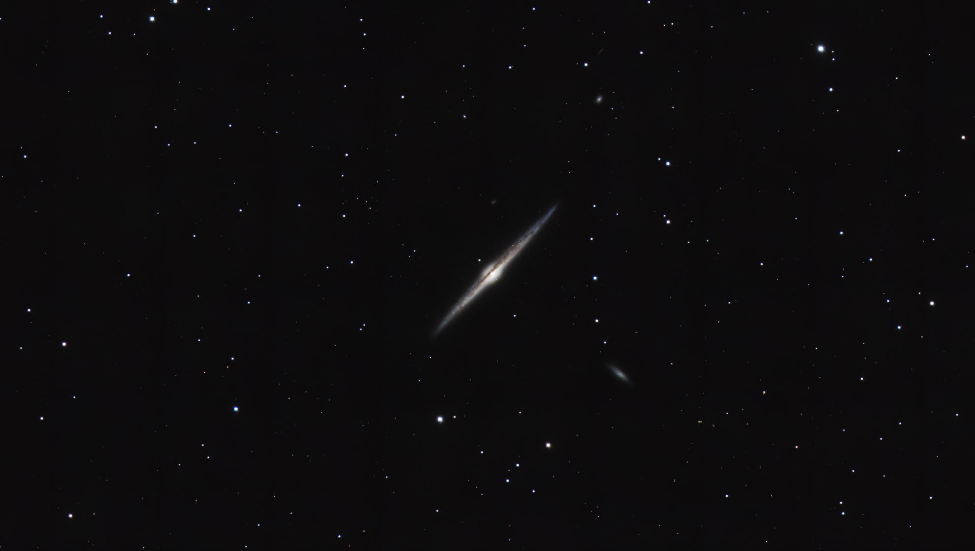 Eine Astrofotografie der Nadel-Galaxie (NGC 4565) vor einem tiefschwarzen, mit fernen Sternen übersäten Hintergrund. Die Galaxie ist exakt von der Seite (Edge-on) zu sehen und erscheint als extrem dünne, spindelförmige Lichtnadel, die diagonal durch das Bild verläuft. In ihrer Mitte ist ein heller, gelblich-weißer galaktischer Kern erkennbar, der von einem markanten, dunklen Staubband in der Längsachse geschnitten wird. In der Umgebung sind zudem zwei deutlich kleinere, diffuse Begleitgalaxien als schwache Lichtflecken sichtbar. Das Bild zeigt eine reduzierte Sternendichte, wodurch die Details der Galaxienstruktur besonders klar hervortreten.